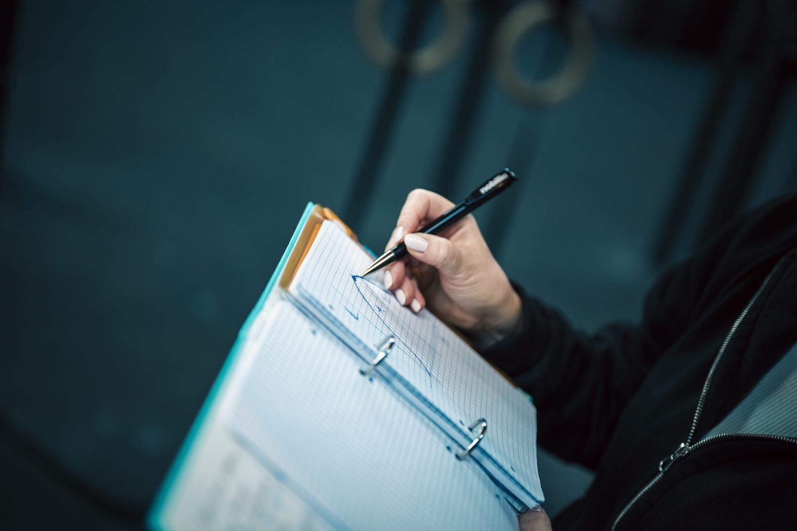 Hand zeichnet mit einem Stift eine Kurve auf Karopapier, im Hintergrund Turnringe